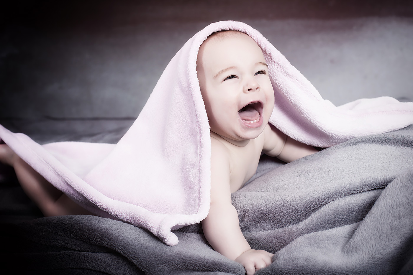 Ein Baby liegt auf einer Decke und schaut lachend unter einem Handtuch hervor